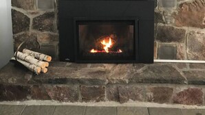 Una chimenea de piedra con fuego encendido, un cuadro colgado encima y una cesta de leña a la izquierda.