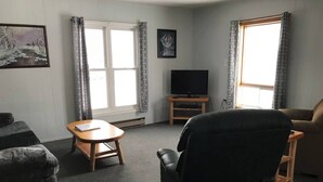 Una sala de estar con un sofá, un sillón y un televisor sobre un soporte de madera. Hay ventanas con cortinas, un cuadro en la pared y una pequeña mesa.