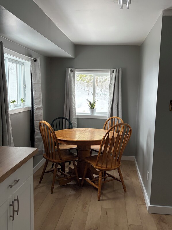 A dining area with a round wooden table and four chairs, a window with curtains, and a view of trees outside.