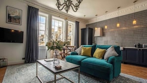 Un salon avec un canapé de couleur turquoise, une table basse et un lustre. Il y a un téléviseur accroché au mur et une fenêtre avec des rideaux.