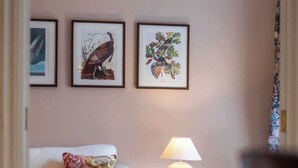 Ein Wohnzimmer mit einem weißen Sofa, einem gemusterten Zierkissen, einem kleinen Beistelltisch mit einer Lampe und gerahmten Kunstwerken an der Wand.