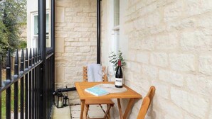 A wooden table with a wine bottle and a book on a balcony with stone walls and a black railing. - Elegant Apartment | Balcony & Free Parking (Bath)