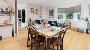 A dining area with a wooden table set for four, surrounded by chairs. There is a green sofa and a window with a view of greenery. - Elegant Apartment | Balcony & Free Parking (Bath)