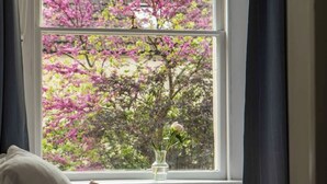 Un dormitorio con cama, almohadas, un jarrón con flores y una ventana que da a árboles rosados.