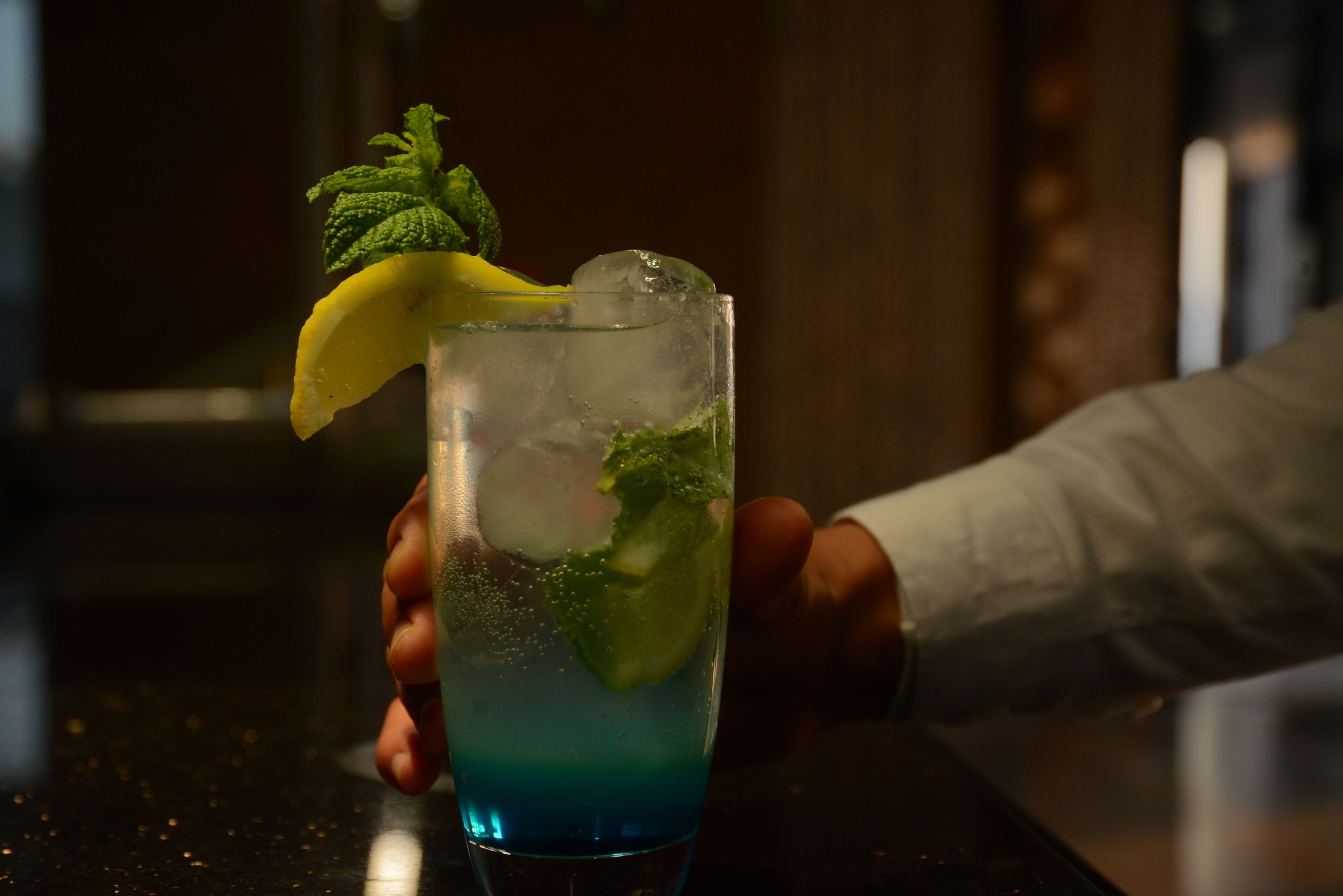 Una persona sosteniendo un vaso con un líquido azul, cubitos de hielo y adornos de lima y menta.