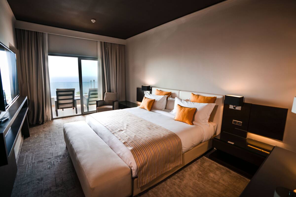 A modern hotel room with a large bed, a TV, a desk, and a balcony with a view of the ocean.