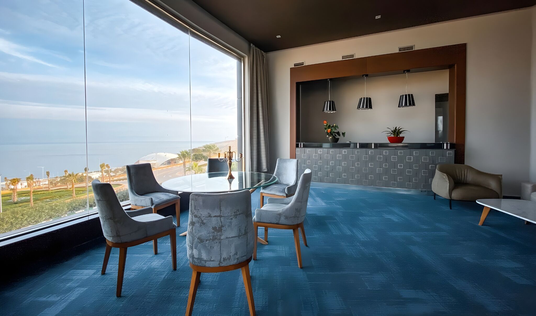 A modern living room with a glass table and chairs, a kitchen area with pendant lights, and a view of the ocean.