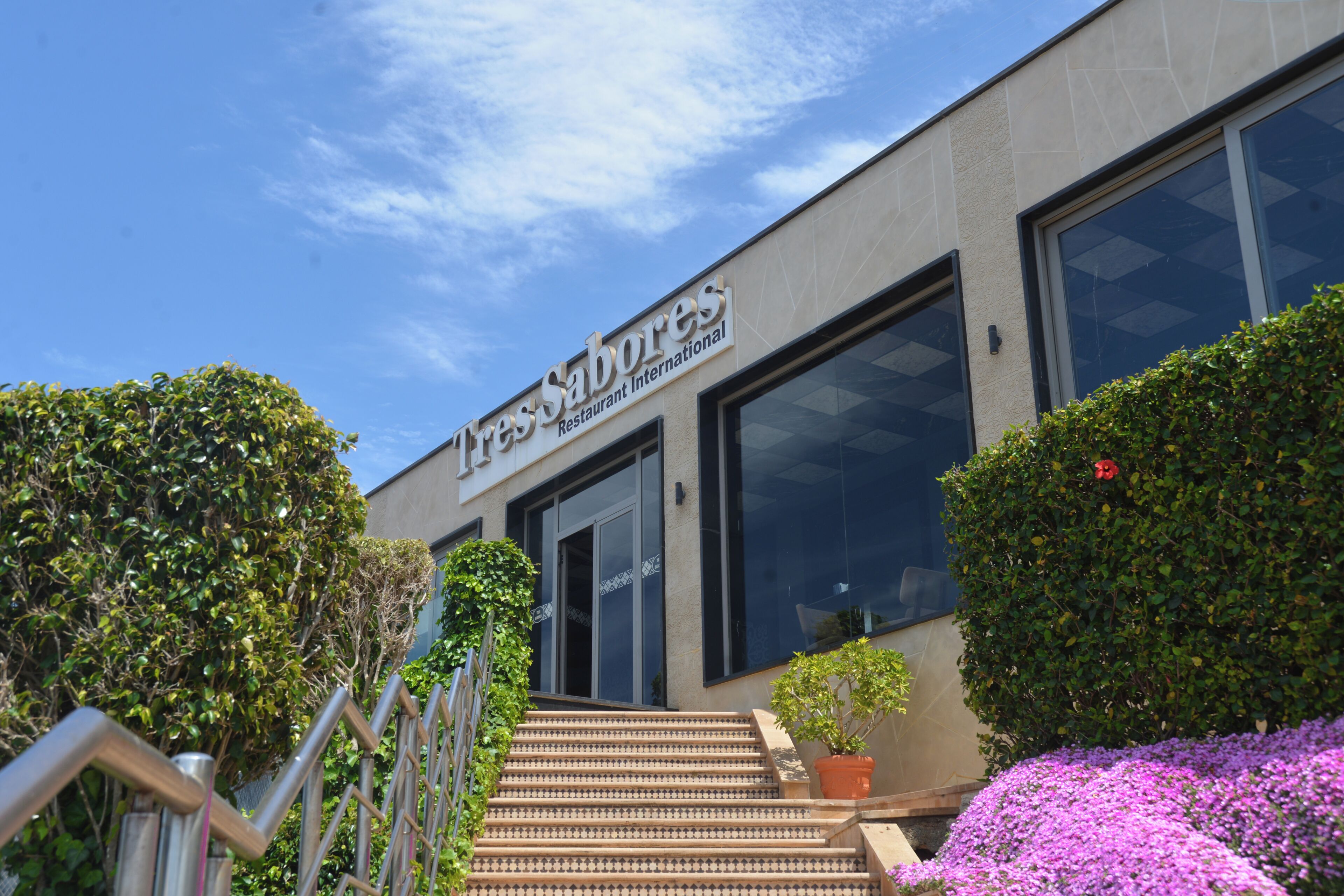 A building with a sign reading 'L'Essentiel Sabores Restaurant International,' a staircase, and surrounding greenery.