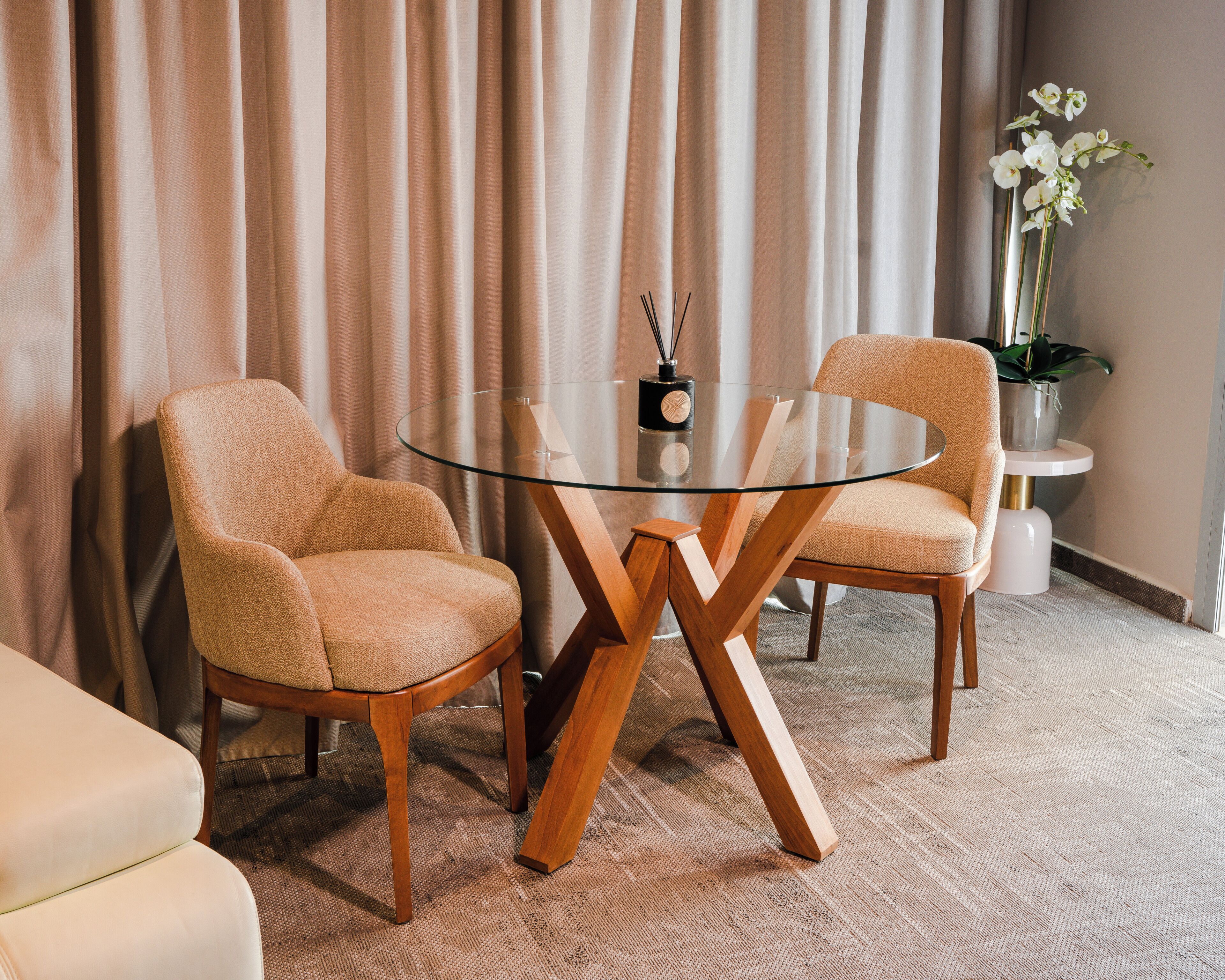 A room with a glass-top table, two upholstered chairs, a potted plant, and a curtain backdrop.