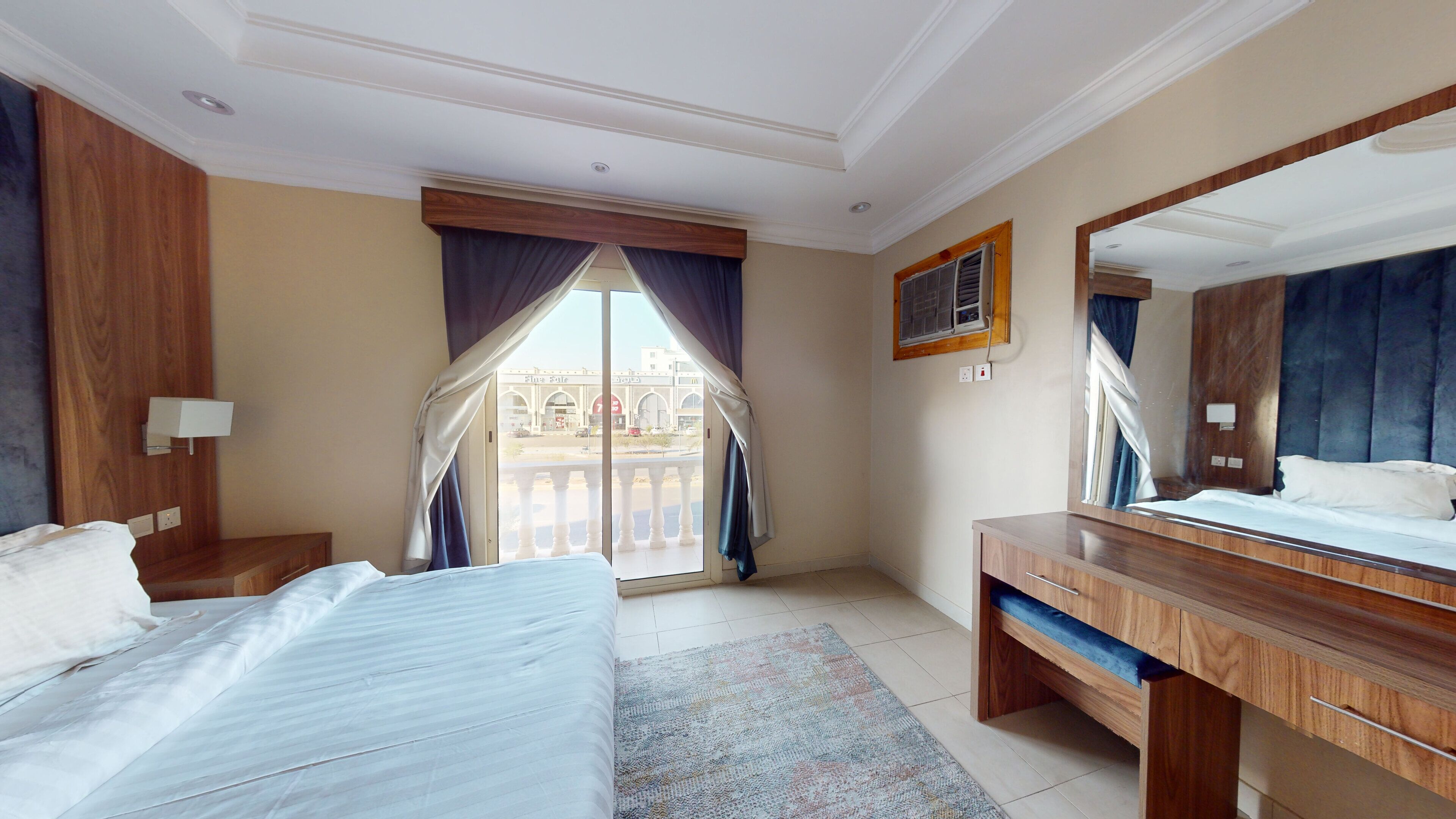 A hotel room with a bed, a desk, a mirror, and a view of a building through the window.