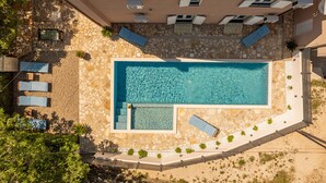 Vista aérea de una piscina con solárium de piedra y azulejos, rodeada de sillas de descanso y un pequeño jardín.