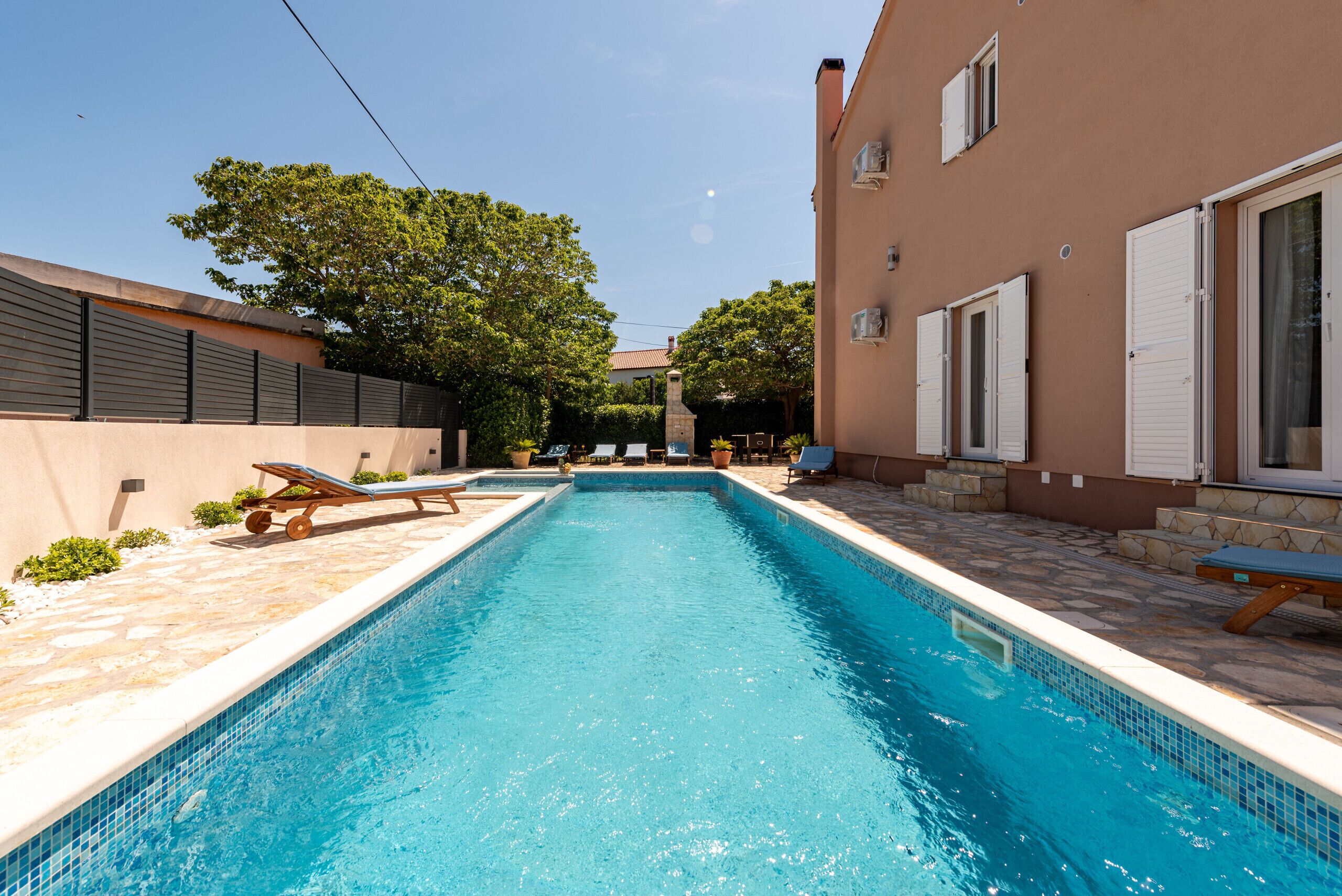 A rectangular swimming pool with a tiled edge, surrounded by lounge chairs and a stone-tiled deck.
