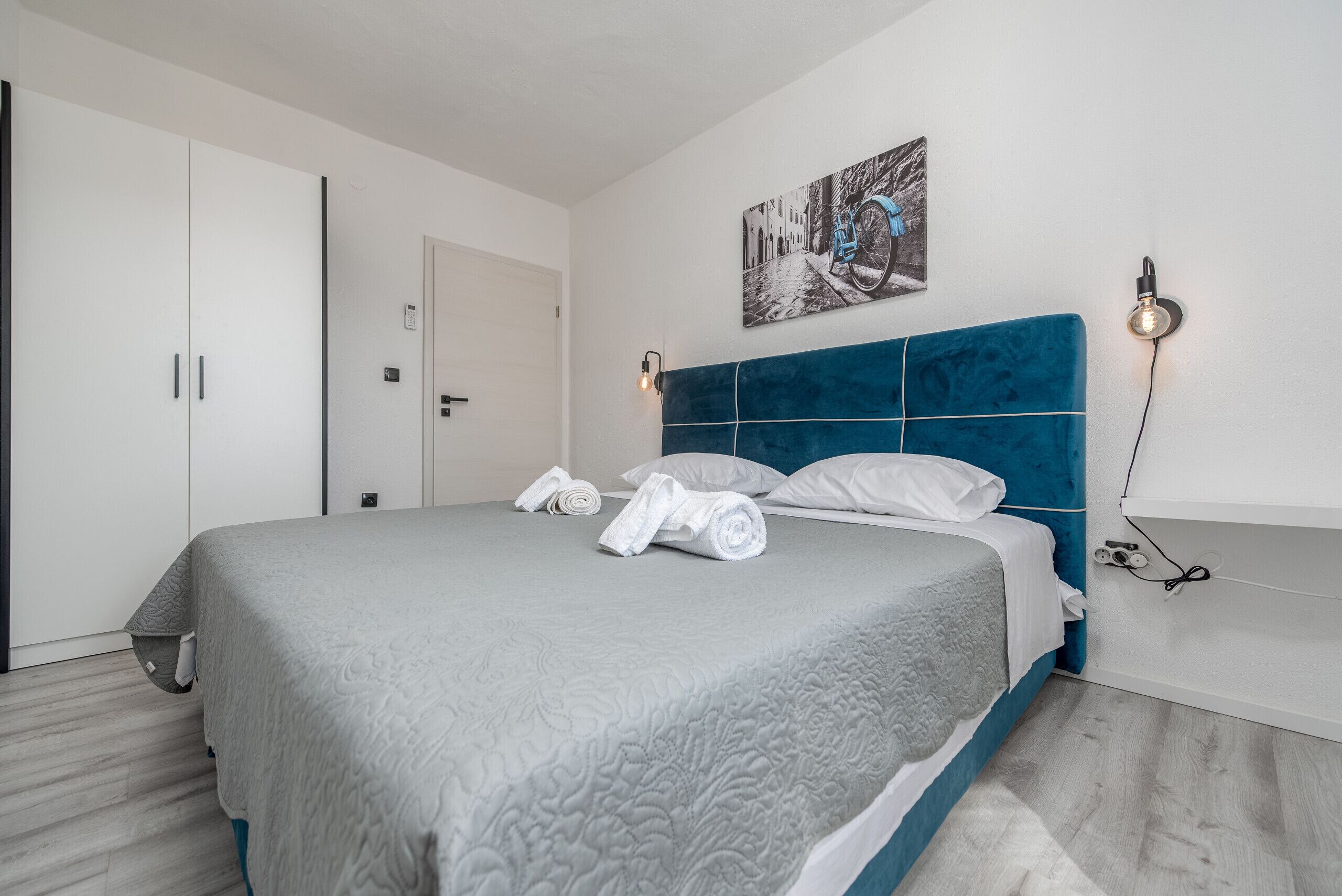 A neatly made bed with a grey quilt, a blue headboard, and a painting of a bicycle on the wall.