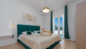 Un dormitorio con una cama tapizada de verde, una mesita de noche con una bandeja, un aire acondicionado de pared y una ventana con cortinas.