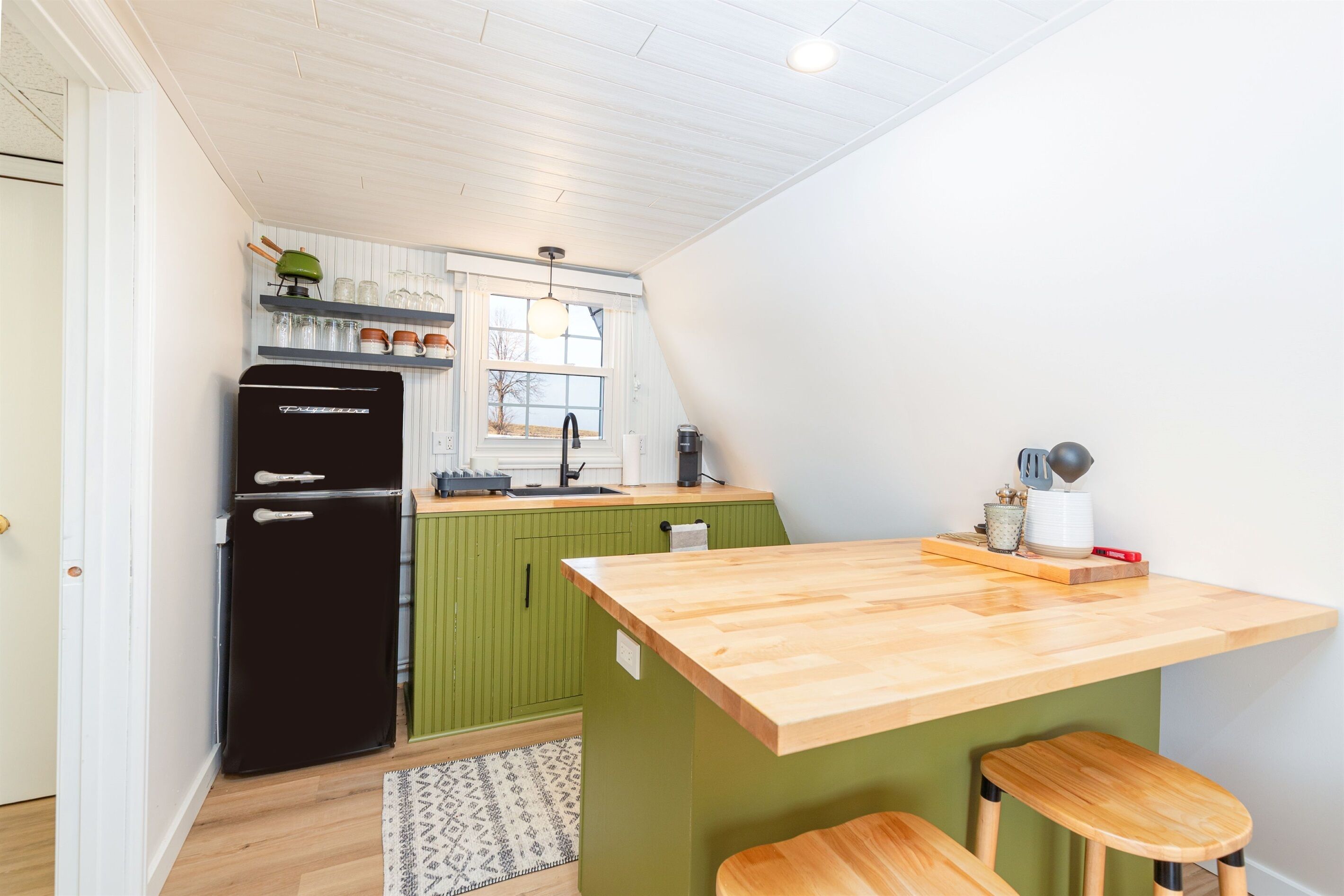 Une cuisine compacte avec un réfrigérateur noir, des armoires vertes, un plan de travail en bois et une fenêtre donnant sur un beau paysage.