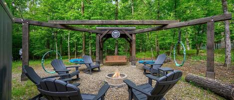 Una pérgola de madera con sillas Adirondack y una chimenea en un área boscosa.