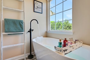 A bathroom with a bathtub, a window with a view of greenery, a towel rack, and a shelf with toiletries. - Arcade Kid Friendly Firepit Grill HotTub (Winston-Salem)