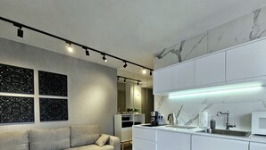Une cuisine moderne avec des meubles blancs, un four à micro-ondes et un évier. Le salon adjacent est équipé d’un canapé avec des coussins.