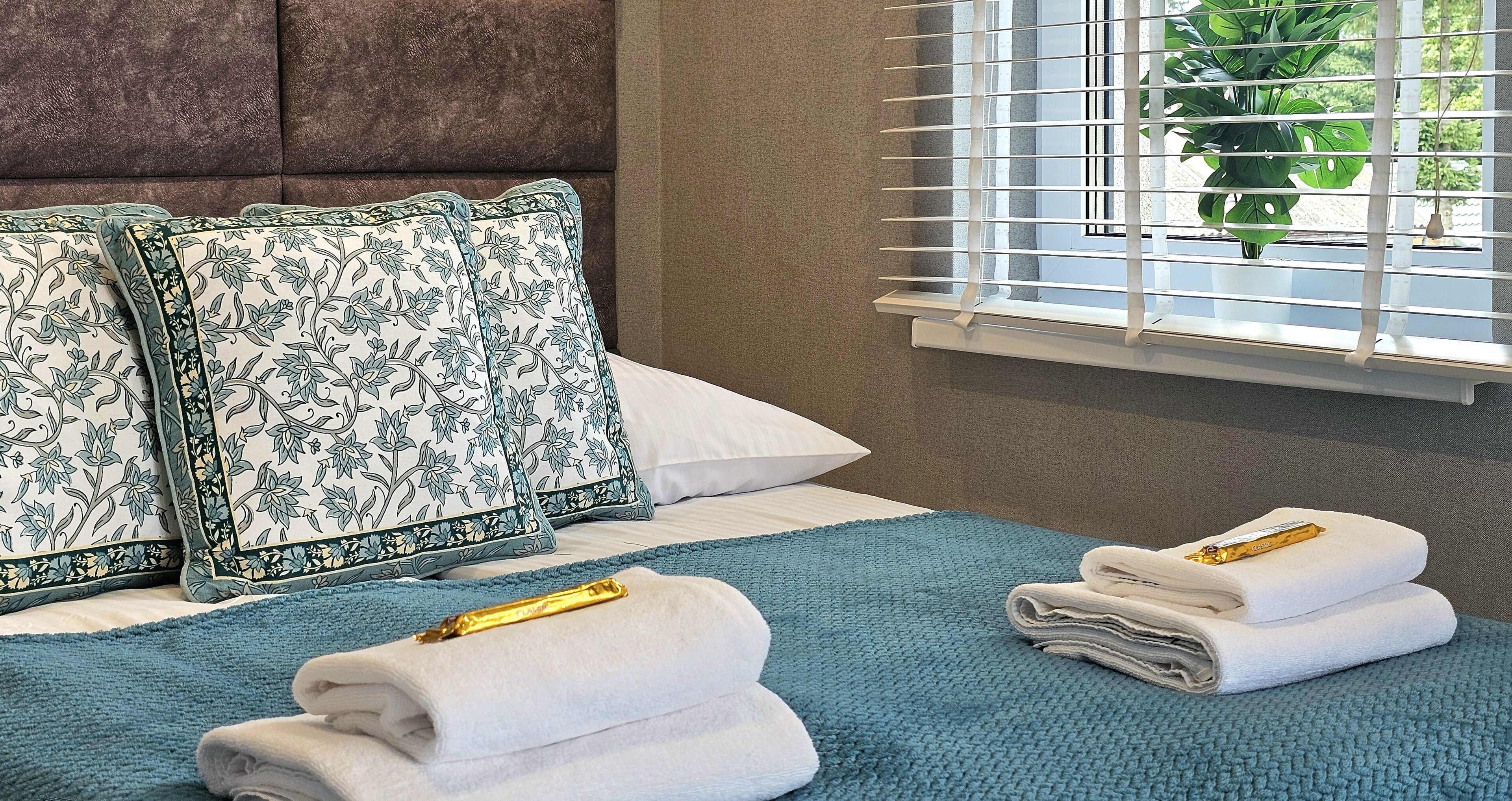 Een net opgemaakt bed met bloempatroon kussens, een blauwe plaid en opgevouwen handdoeken met gouden handvat.