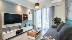 A modern living room with a flat-screen TV, a grey sofa, a wooden coffee table, and a large window with curtains. - Mediteranean Luxury Apartment (Chania)
