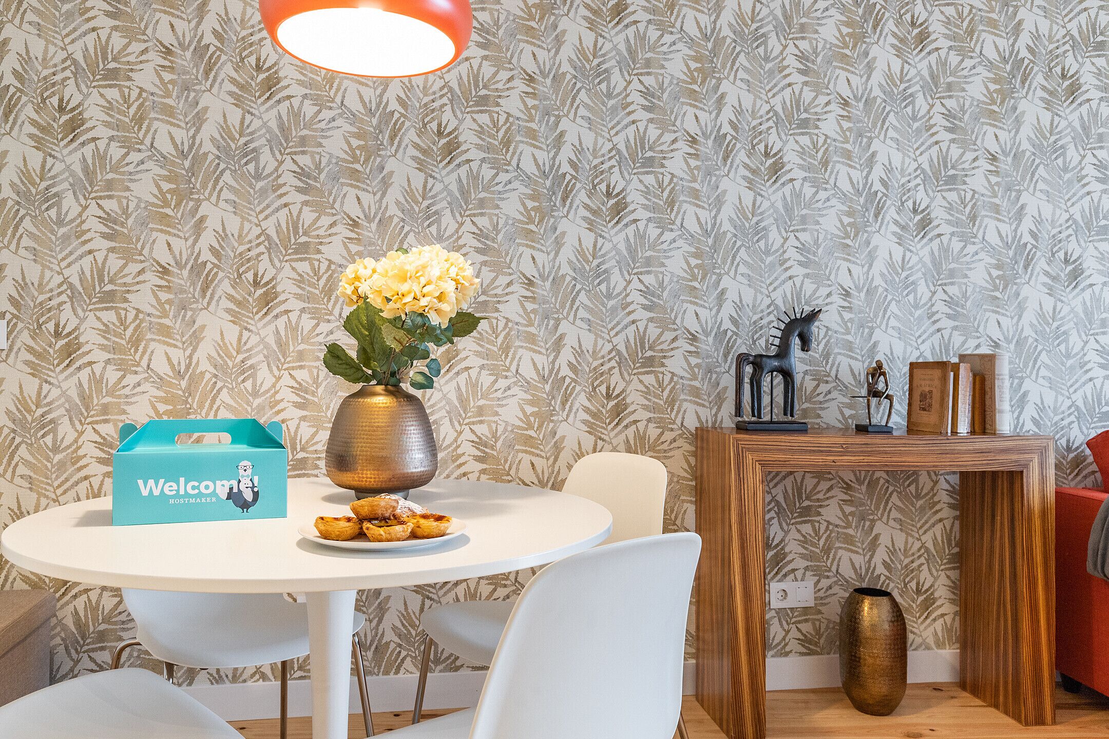 Uma área de jantar com uma mesa branca redonda, um vaso com flores amarelas, uma caixa de boas-vindas e uma estátua decorativa de um cavalo.