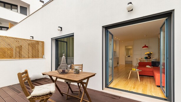 Eine Holzterrasse mit Tisch und St ühlen, eine Glastür, die in einen Wohnbereich mit einem roten Sofa und einer Küche im Hintergrund führt.