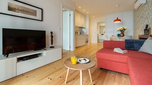 Ein modernes Wohnzimmer mit einem Flachbildfernseher, einem roten Sofa, einem kleinen Holztisch mit einem Glas Orangensaft und einer Küche im Hintergrund.