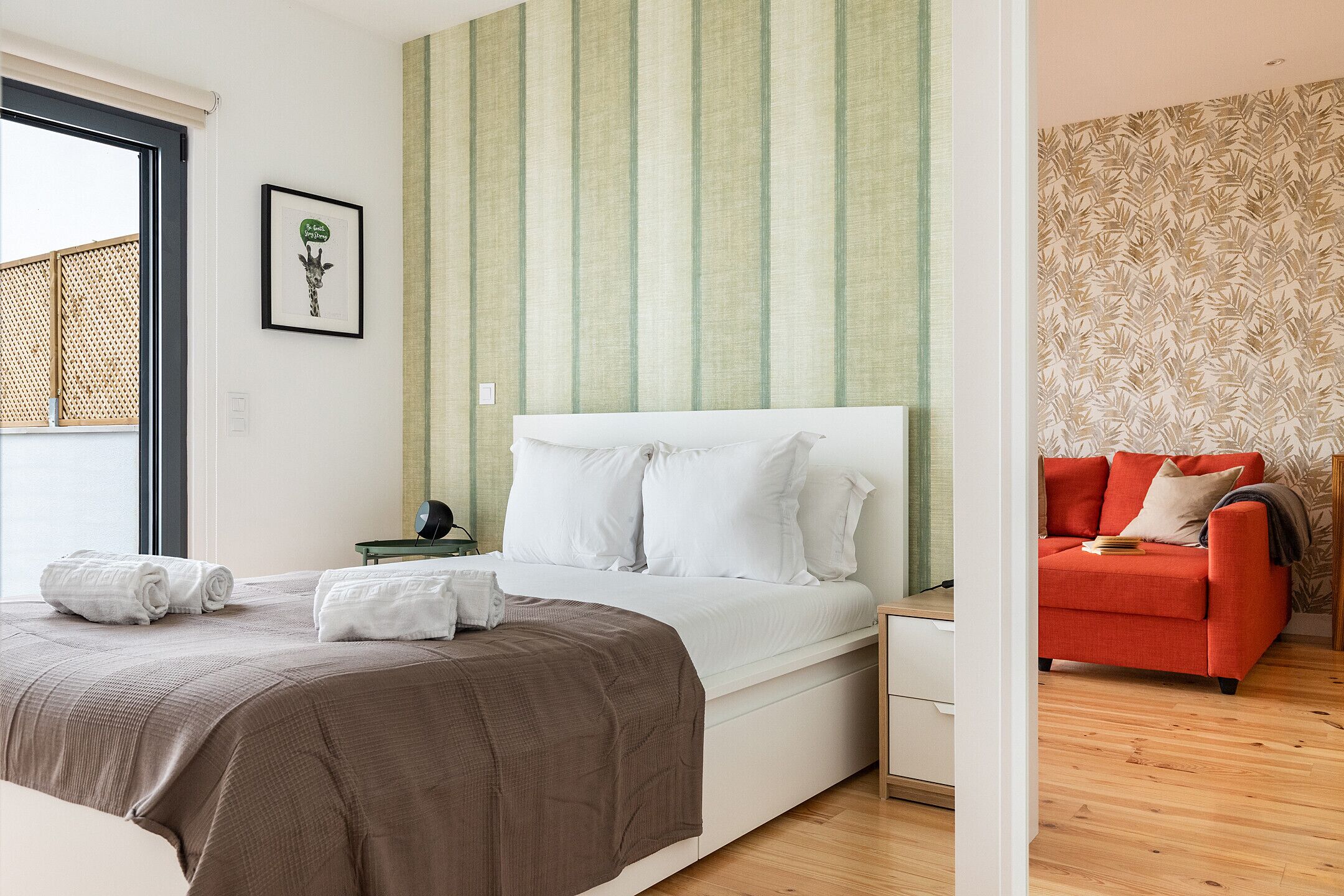 A bedroom with a bed, a nightstand, a red sofa, and a window with a view.