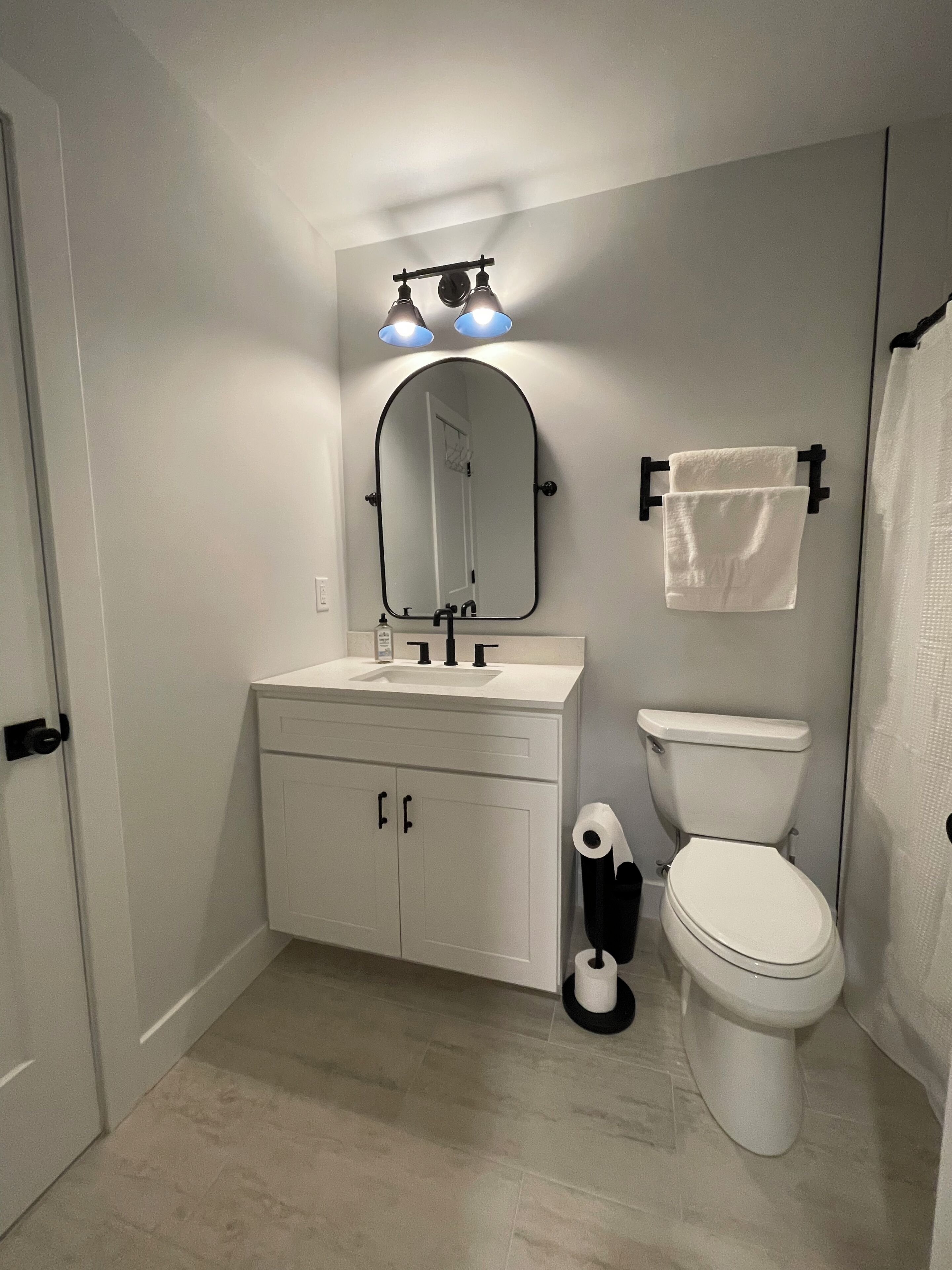 Ein Badezimmer mit einem weißen Waschbecken, einem Spiegel und einer Toilette. Zwei Leuchten befinden sich über dem Spiegel, und ein Handtuchhalter mit einem Handtuch ist vorhanden.