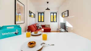 A modern living room with a white table, a red sofa, and a TV. There are framed pictures on the wall, a blue 'Welcome' box, and a glass of orange juice on the table. - Belem Design 08 by Homing (Lisbon)