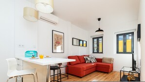 A modern living space with a white table, red sofa, and a glass coffee table. There is a framed artwork on the wall and a window with yellow blinds. - Belem Design 08 by Homing (Lisbon)