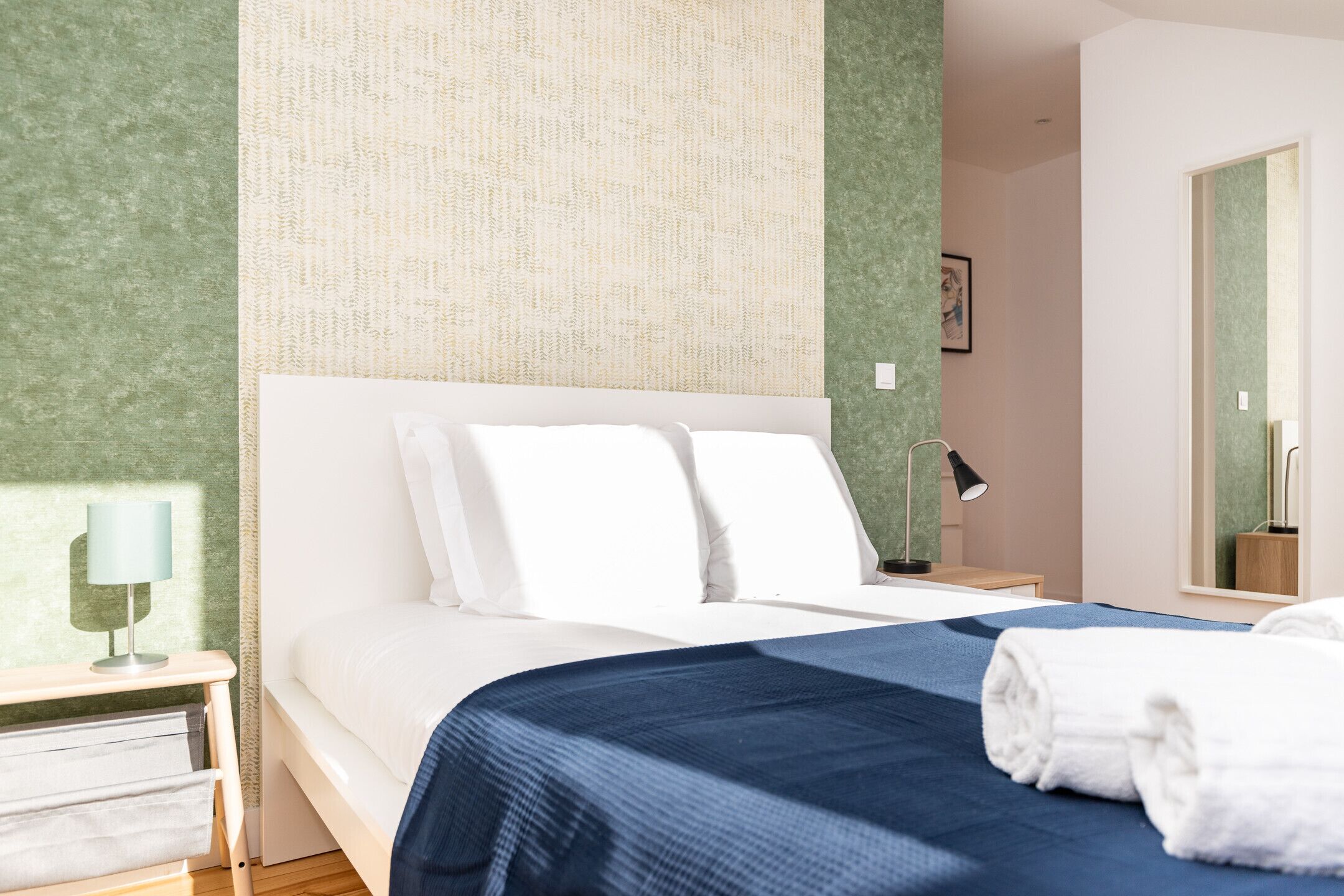 A neatly made bed with white linens and a blue bedspread, a bedside lamp, and a mirror on the wall.