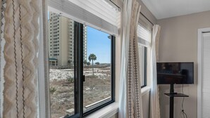 A room with a large window, a television on a stand, and a view of a building and palm tree outside. - Sugar Beach C-11 (Navarre)