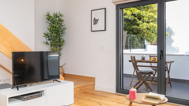 Un moderno salón con un televisor de pantalla plana sobre un soporte blanco, una planta en maceta, un cuadro en la pared y un ventanal corredero que da a un área exterior con una mesa y sillas.