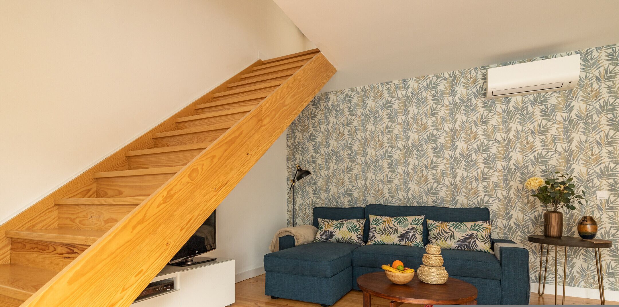 Un salón estilo loft con una escalera de madera, un conjunto de sofá azul, una pequeña mesa de madera con fruta y un papel tapiz estampado.