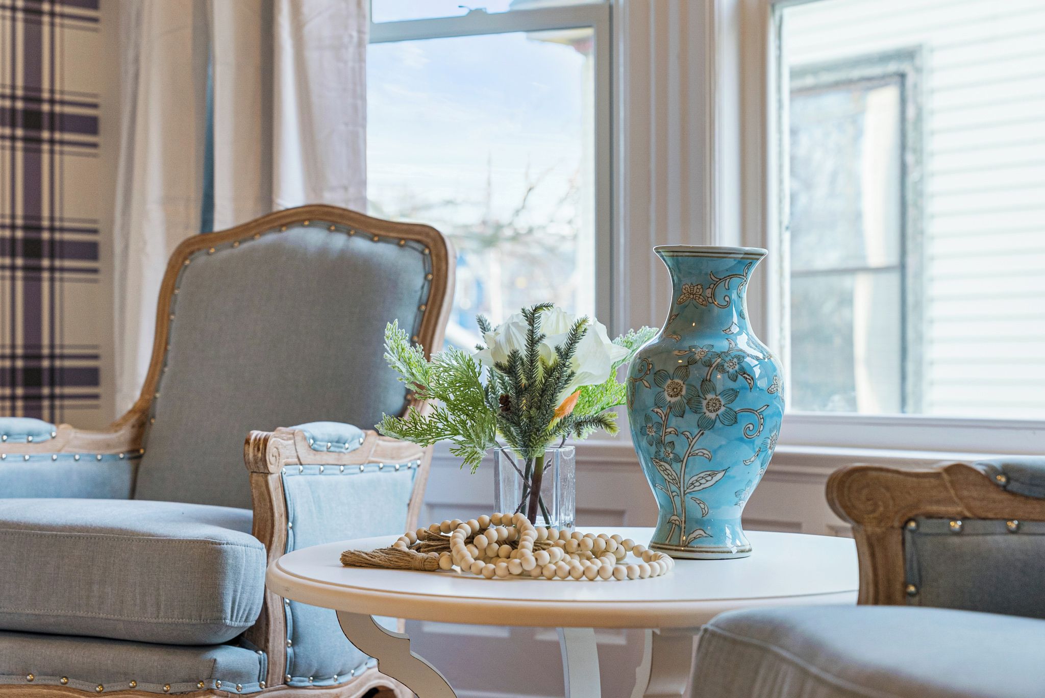 Una habitación con un sillón azul, una mesita con un florero y una guirnalda de cuentas, y una ventana con cortinas.