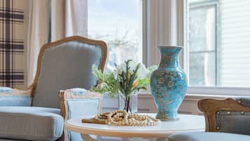 Una habitación con un sillón azul, una mesita con un florero y una guirnalda de cuentas, y una ventana con cortinas.