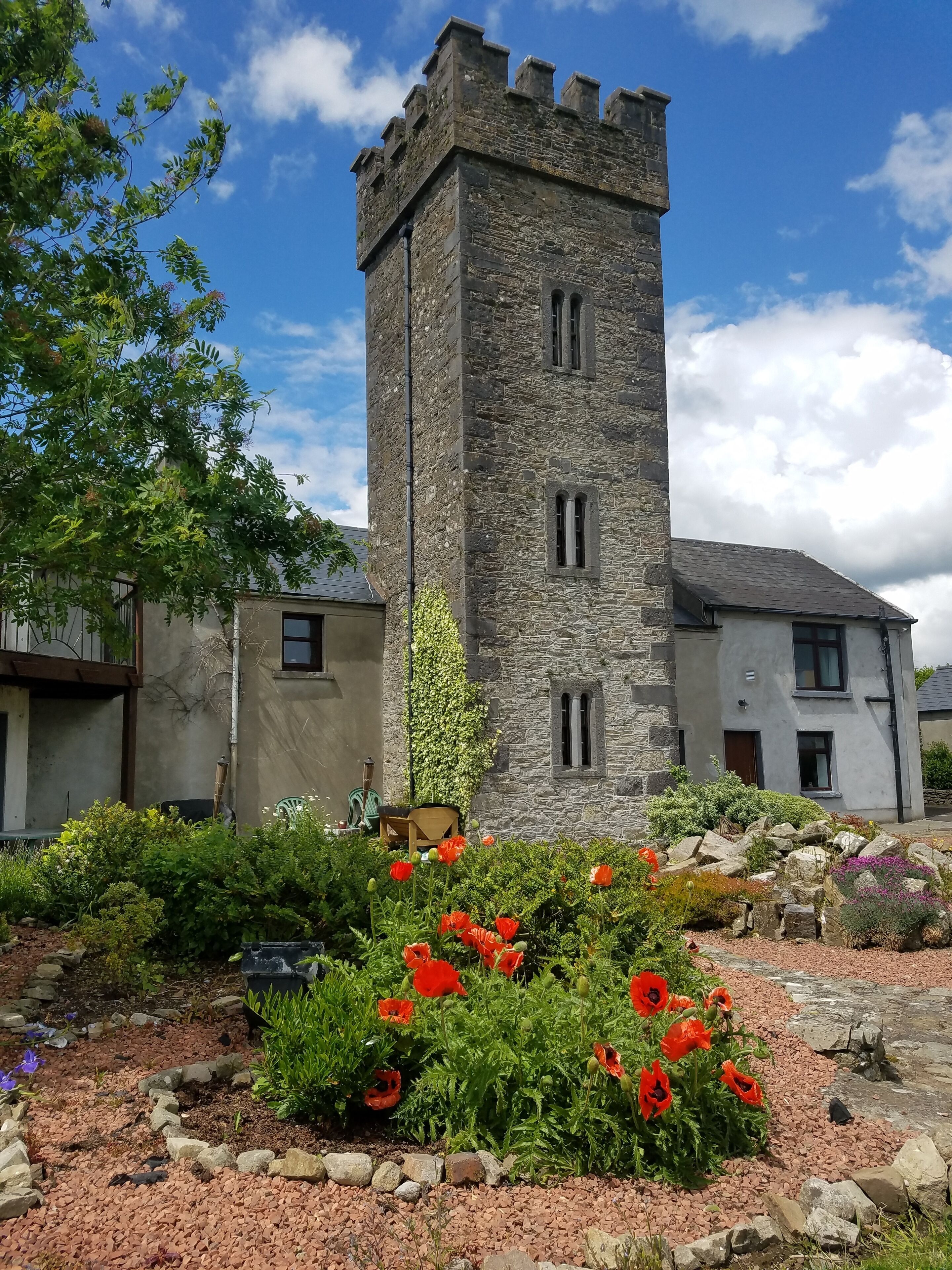 Ein Steinturm mit einem Turmzimmer, umgeben von einem Garten mit roten Klatschmohnblumen und verschiedenen Sträuchern.