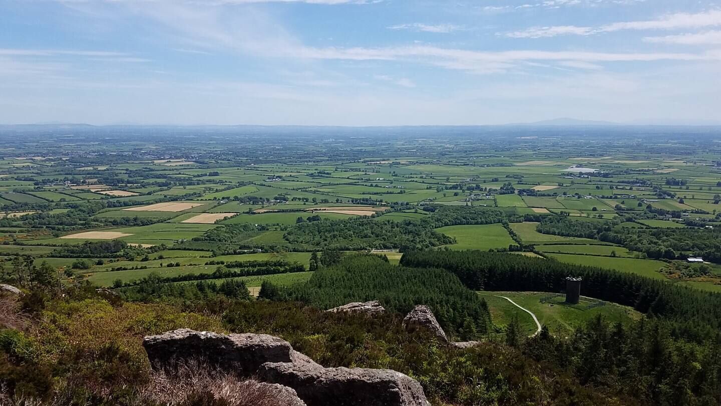 Une vue panoramique d’un paysage rural, avec des champs vallonnés, une route sinueuse et une chaîne de montagnes lointaine.