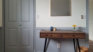 Une pièce avec une porte fermée, un miroir fixé au mur, une console en bois sur laquelle est posé un vase de fleurs, et un panier en osier.