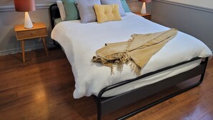 Un lit bien fait, recouvert d’une couette blanche et d’un plaid beige, flanqué de deux tables de chevet avec des lampes, dans une pièce au plancher en bois.