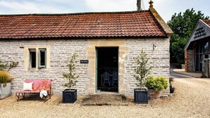 Un bâtiment en pierre avec un toit recouvert de tuiles rouges, un banc et deux plantes en pot devant.