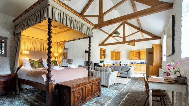 Un lit à baldaquin à quatre poteaux avec une tête de lit en bois, un sol imitant la pierre, une table à manger en bois avec des chaises, et une cuisine en arrière-plan.