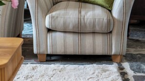 Un fauteuil à rayures avec un coussin vert, une table d’appoint en bois et un tapis blanc dans une pièce dont le sol est recouvert de pierres.