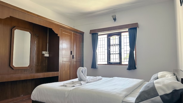 A hotel room with a large bed, a wooden wardrobe, a window with curtains, and a wall-mounted light fixture. - Travel & Stay (Ootacamund)