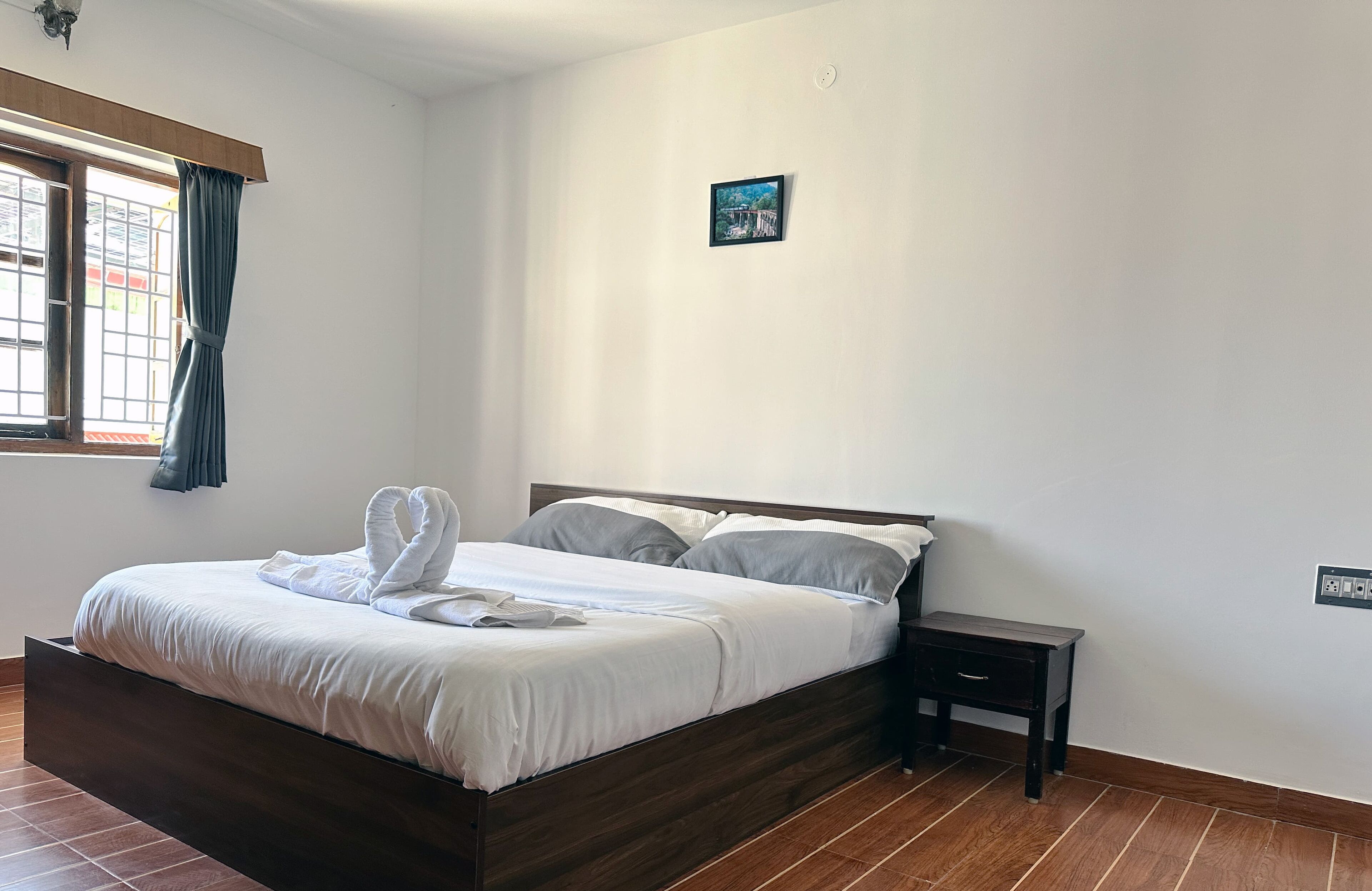 A neatly made bed with white linens and two pillows, a small bedside table, and a window with curtains.