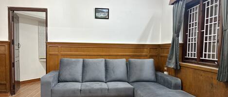 Un sofá seccional gris en una habitación con paneles de madera, un cuadro enmarcado en la pared y una ventana con cortinas.