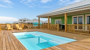 En træterrasse med en pool, udendørs stole og en bygning med grøn facade og hvide vinduer.