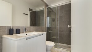 Un baño moderno con un lavabo blanco, dos dispensadores de jabón negros, un inodoro y una zona de ducha con mampara de vidrio.