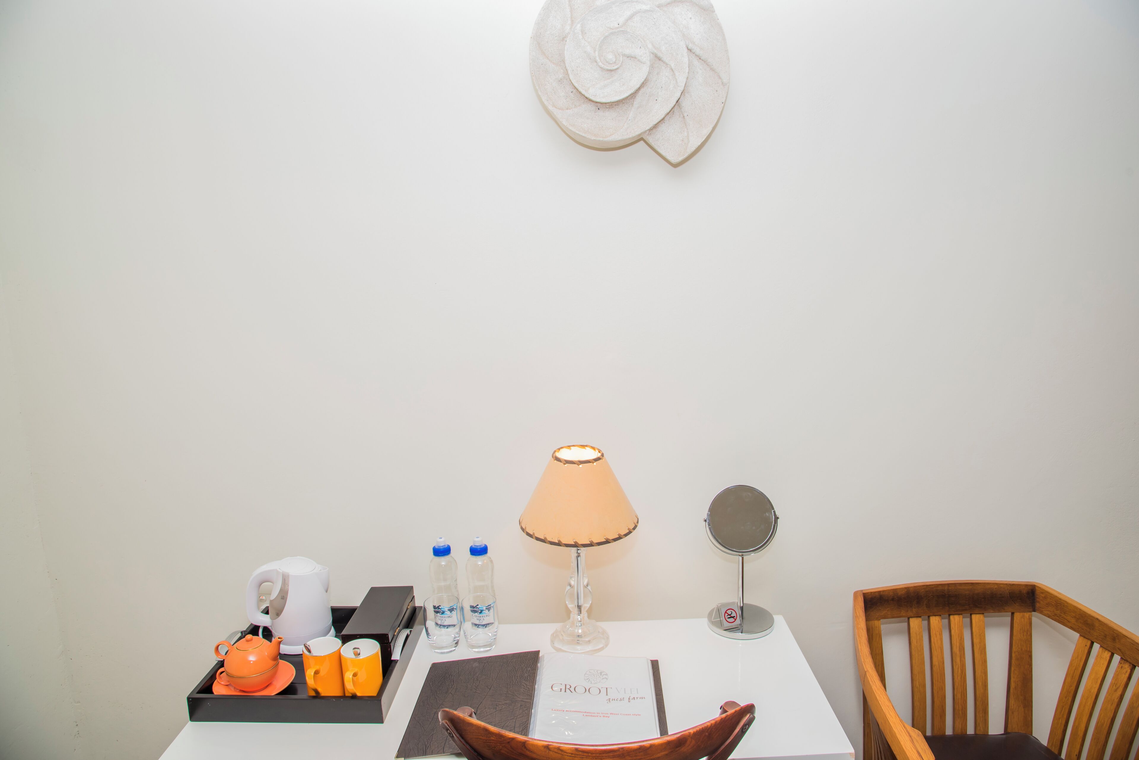 A hotel room with a white table, a wooden chair, a decorative wall piece, a tray with a teapot, cups, and water bottles.